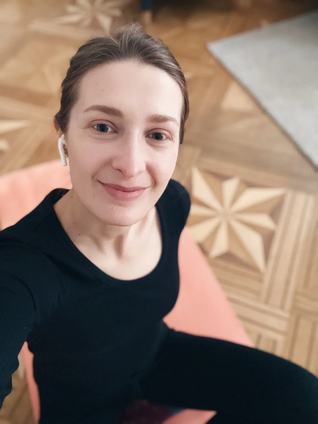 Žena v černém sportovním topu sedí na broskvově růžové podložce na parketách se sluchátky.
