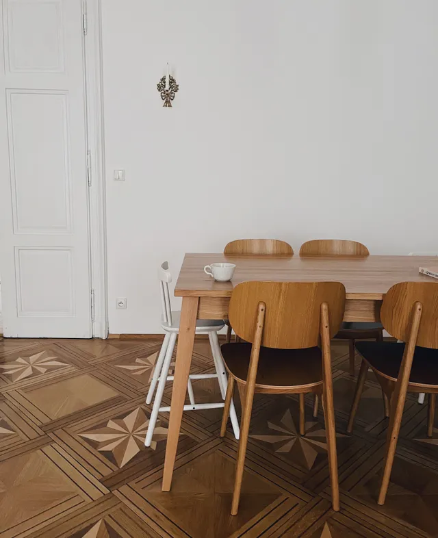 Jídelní kout s dřevěným stolem, židlemi a bílou jídelní židličkou na parketách s hvězdovým vzorem.