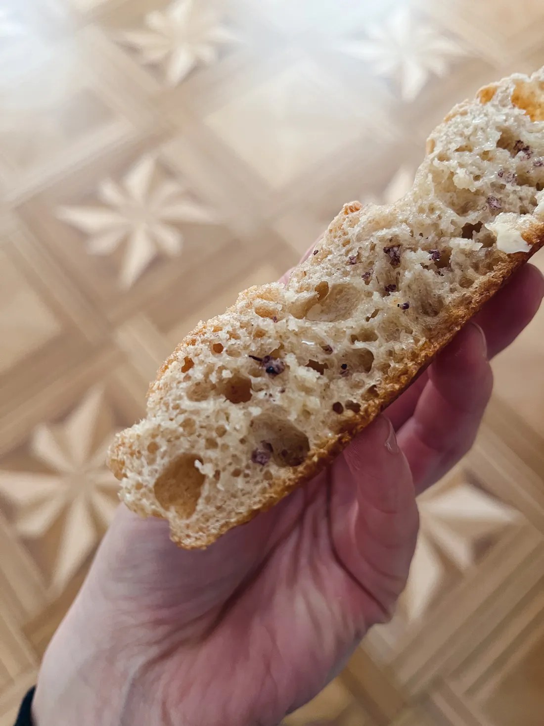 Řez křupavé fokáče s vzdušnou střídkou a tmavými semínky držený v dlani nad parketami.