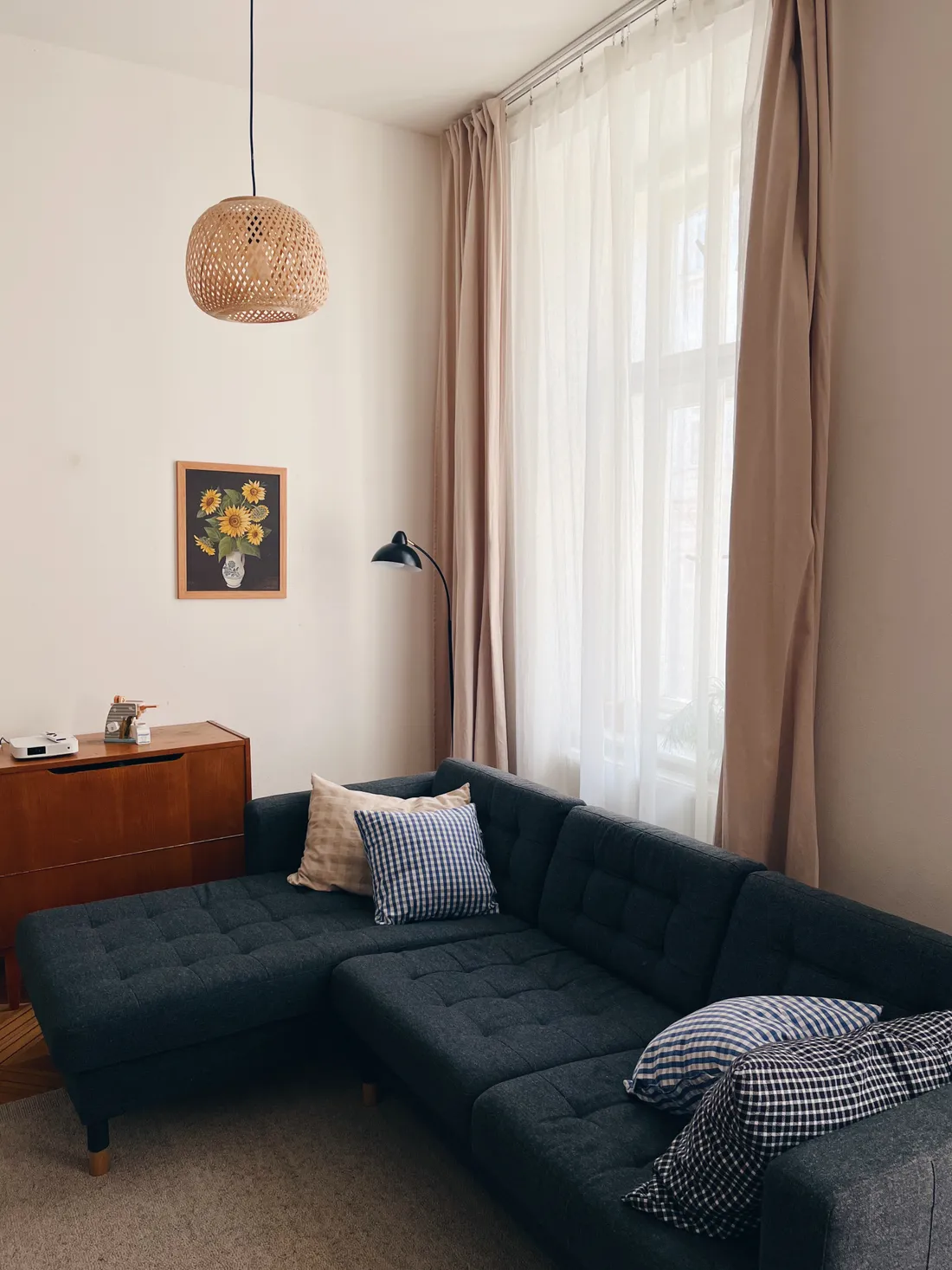Tmavě šedá rohová pohovka s polštáři, proutěným závěsným světlem a obrazem slunečnic u okna.